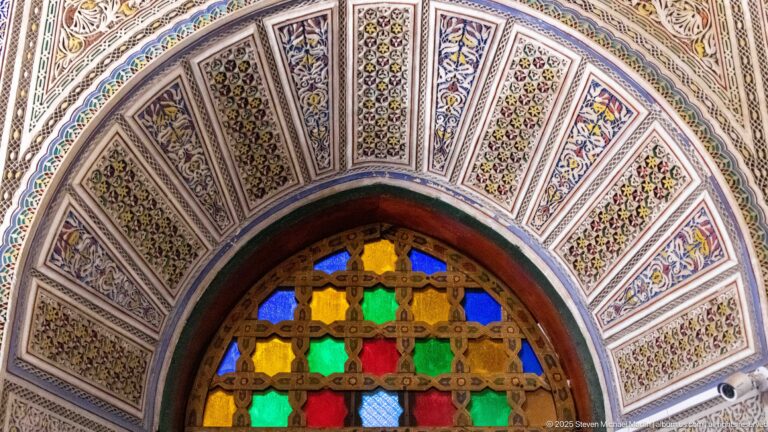 Close view of an arched colored-glass window surrounded by radiating painted floral and geometric panels.