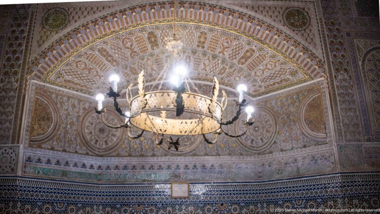 Large chandelier in front of an ornate wall with circular medallions, dense patterning, and blue geometric zellij below.