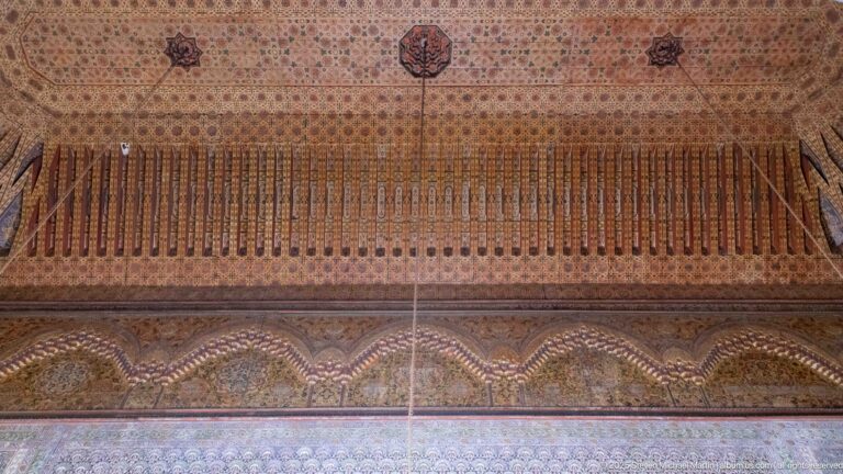 Close upward view of a decorated wooden ceiling edge with vertical painted elements and a scalloped stalactite-like band.