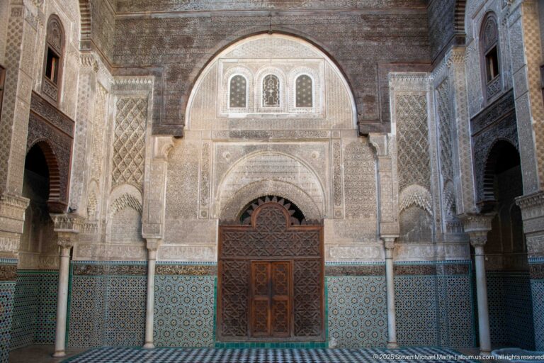 Frontal Attarine Madrasa courtyard façade with carved wooden doorway, stucco arches, three upper windows, columns, and zellij.