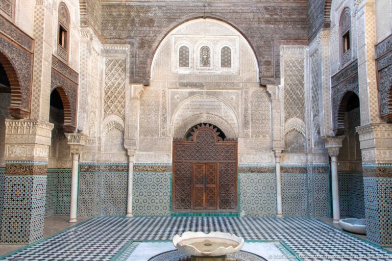 Attarine Madrasa courtyard with shallow central marble basin, carved wooden doorway, stucco façade, zellij, and upper windows.