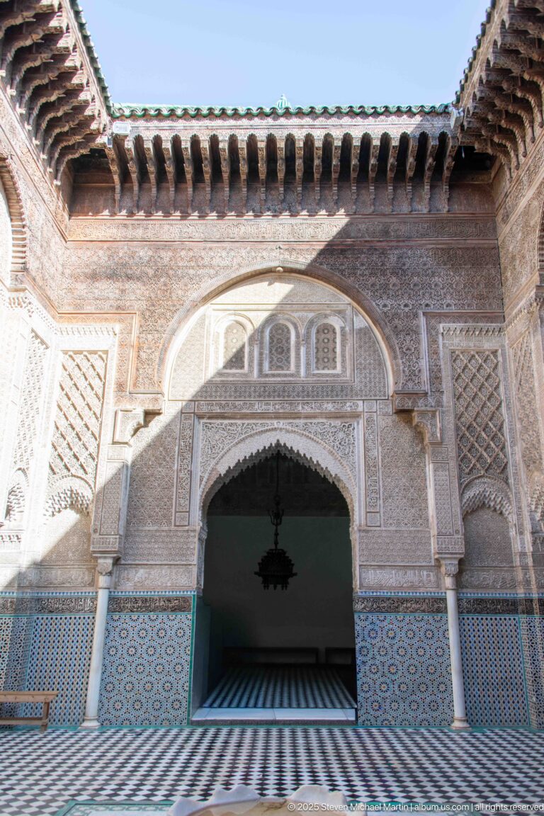 Wide Attarine Madrasa courtyard view with monumental arch, carved cedar eaves, zellij walls, columns, and dark interior opening.
