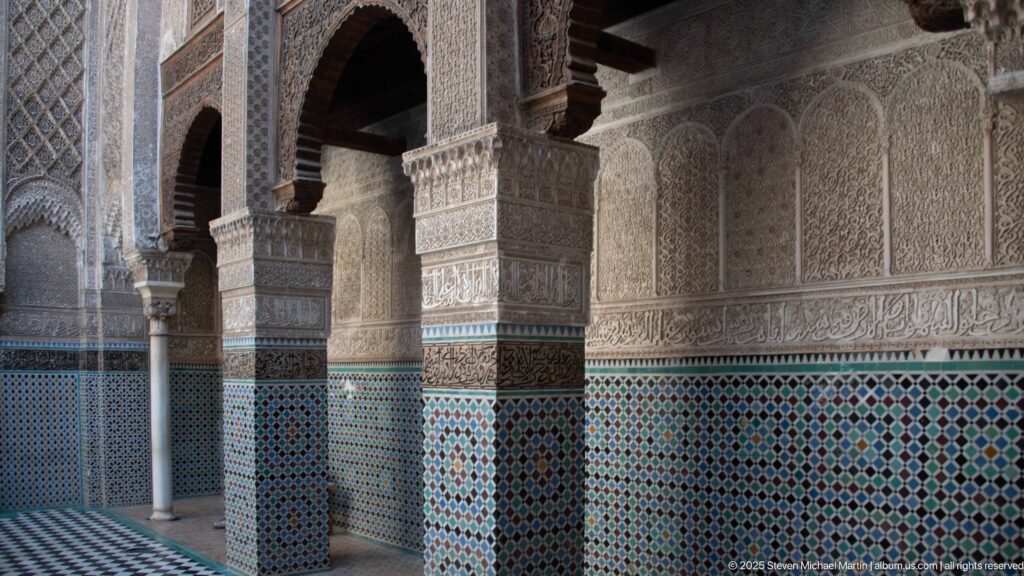 Oblique Attarine Madrasa arcade with carved stucco columns, patterned zellij, calligraphic bands, and shadowed arches.