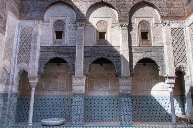 Two-story Attarine Madrasa courtyard façade with lower arches, small upper windows, columns, zellij panels, and marble basin.