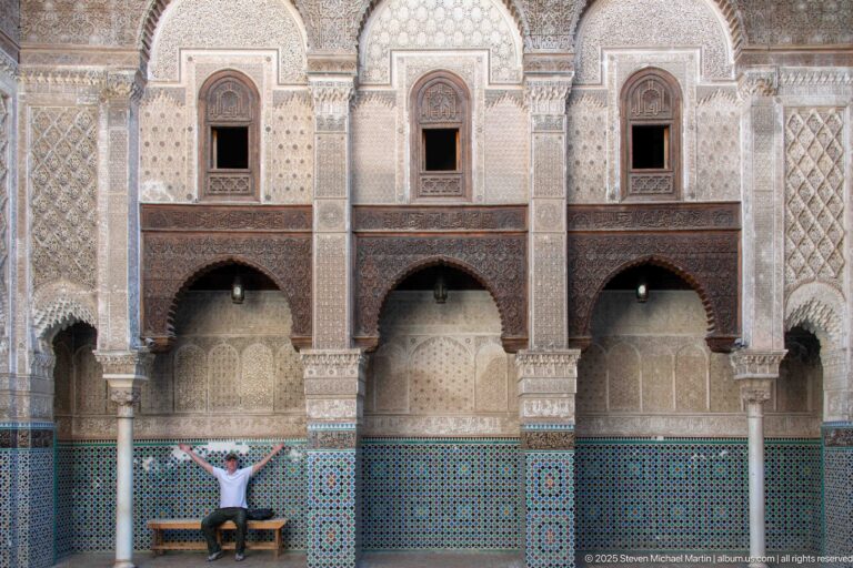 Seated visitor beneath two-story Attarine Madrasa courtyard arches with upper windows, carved stucco, columns, and zellij.