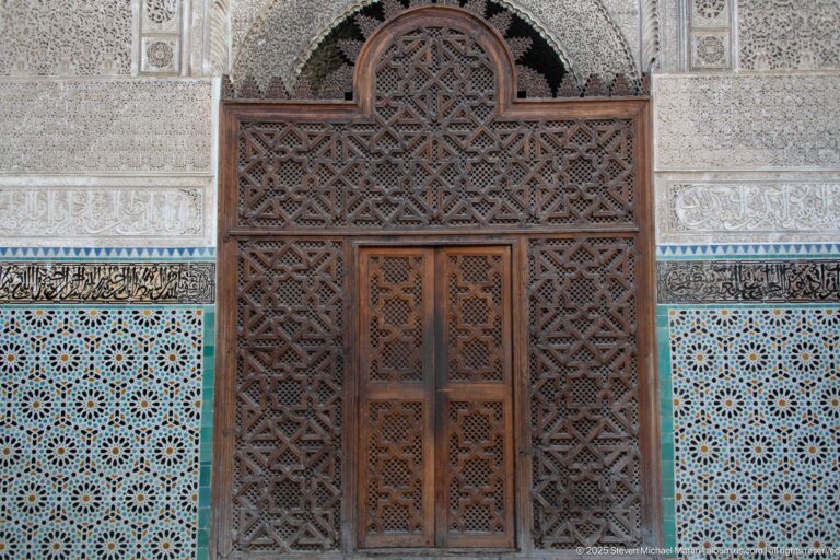 Carved cedar screen and double door with interlaced star patterns, framed by carved stucco and blue-green zellij.