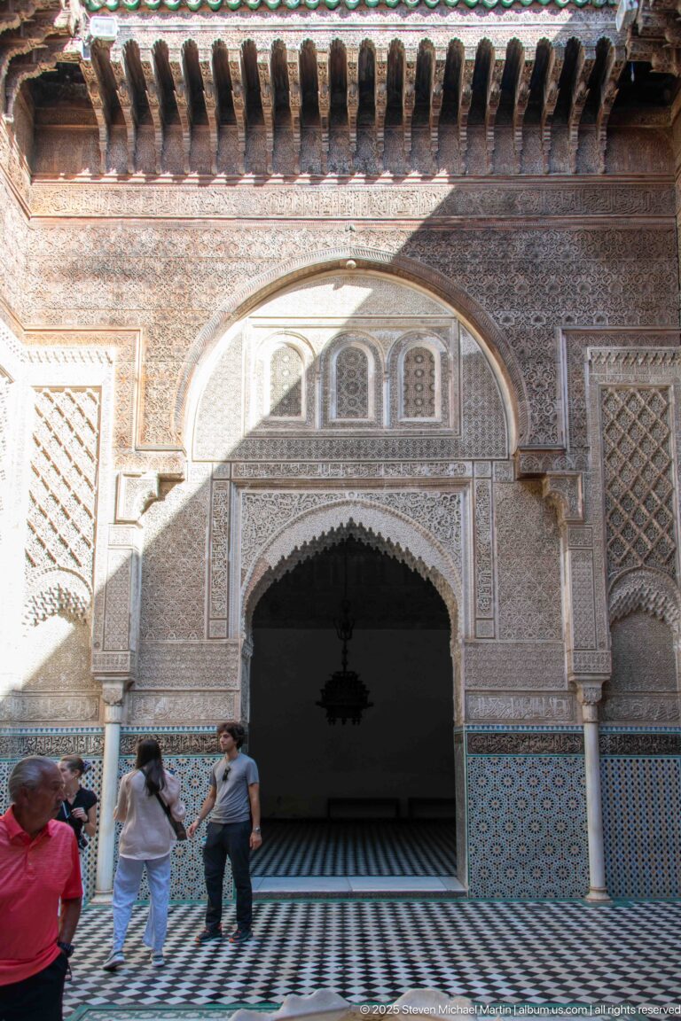 Main Attarine Madrasa courtyard façade with horseshoe arch, carved stucco, cedar wood, zellij, tiled floor, and visitors.