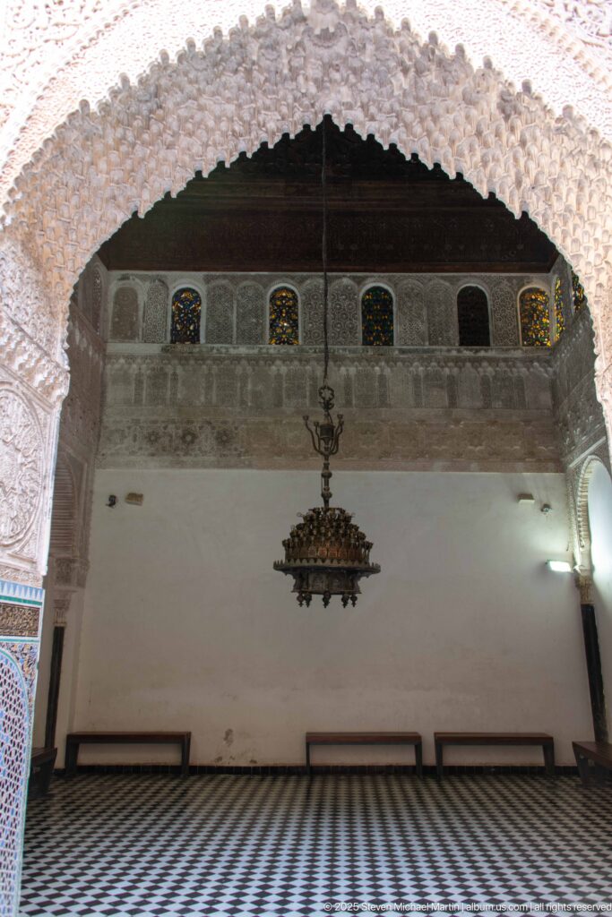 Prayer hall seen through a carved scalloped arch, with hanging chandelier, small stained-glass windows, benches, and tiled floor.
