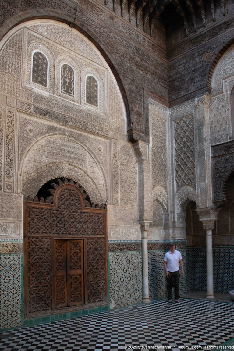 Angled view of Attarine Madrasa carved wooden doorway, stucco arch, zellij walls, columns, and seated visitor.