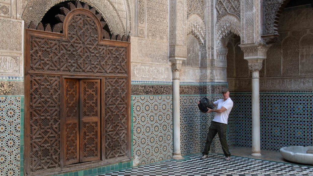 Visitor standing beside the carved cedar doorway, columns, zellij wall, and black-and-white courtyard pavement.