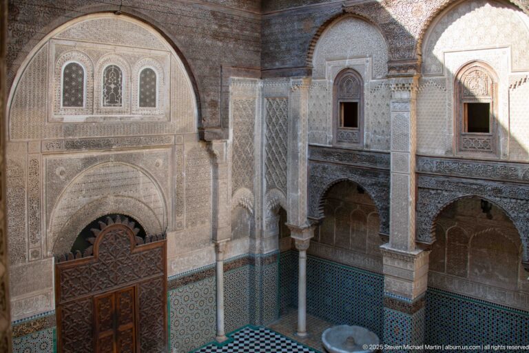 Elevated diagonal view across the Attarine Madrasa courtyard with main doorway, side arcade, upper windows, zellij, and basin.
