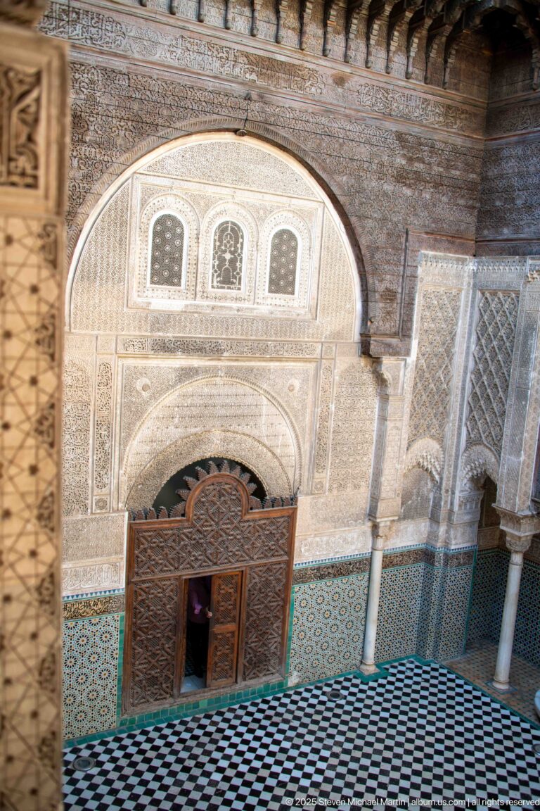 Upper-level view down across black-and-white courtyard tiles toward the carved wooden doorway and zellij façade.