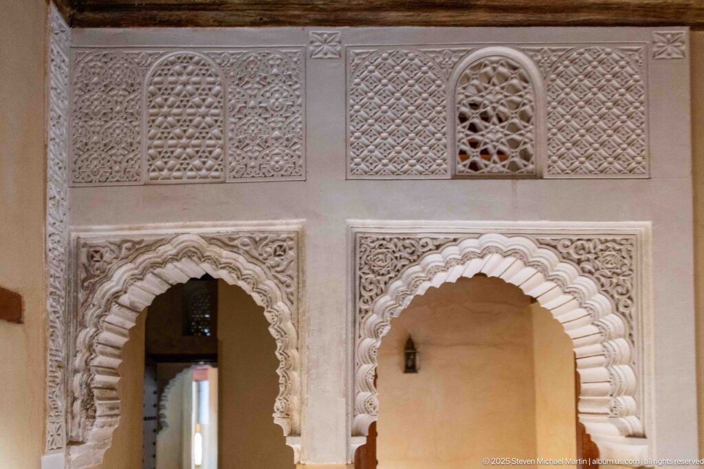 Two scalloped carved-stucco arches with geometric panels and a small lattice opening in the Attarine Madrasa upper level.