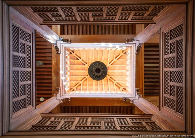 Upward view of cedar beams and lattice panels framing a bright central roof opening with a circular hanging fixture.
