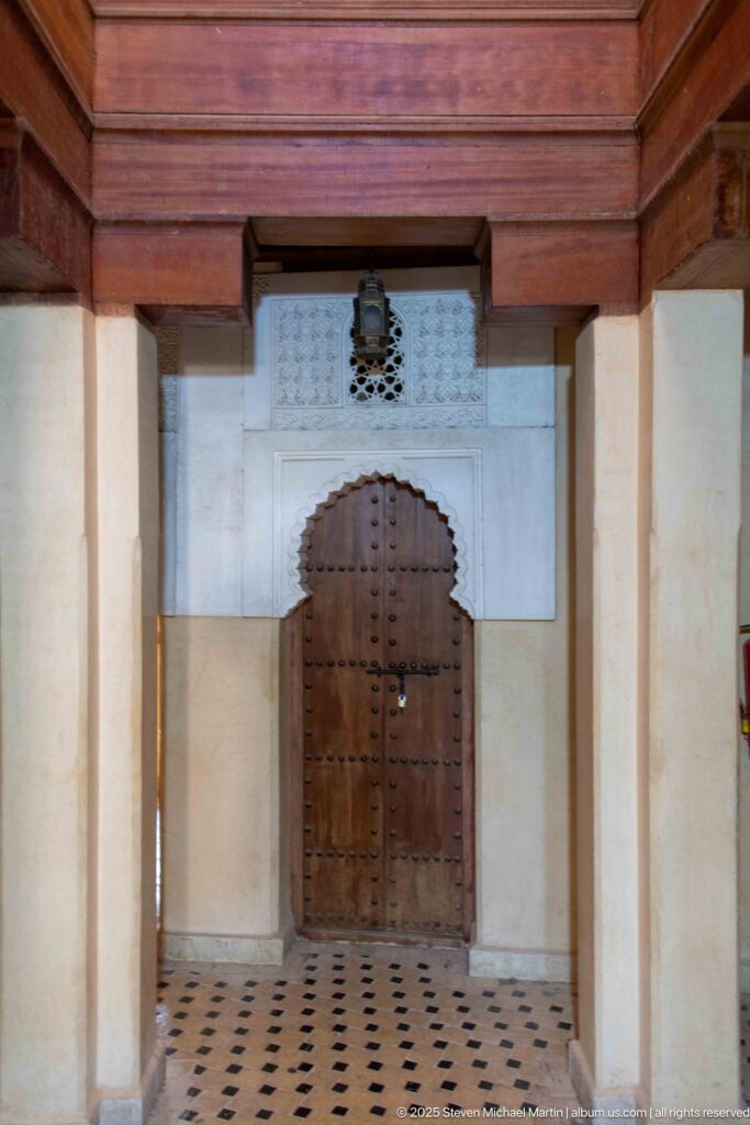 Studded wooden door beneath heavy cedar beams in a plain upper corridor with patterned tile floor.