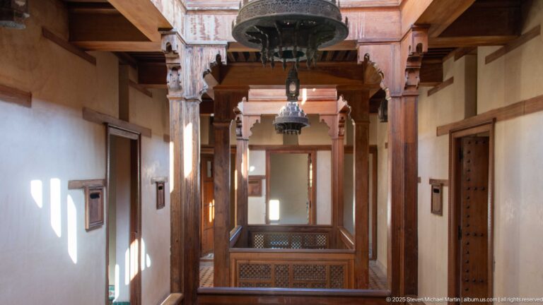 Attarine Madrasa upper wooden gallery with carved posts, railings, hanging lamps, small doors, and pale plaster walls.