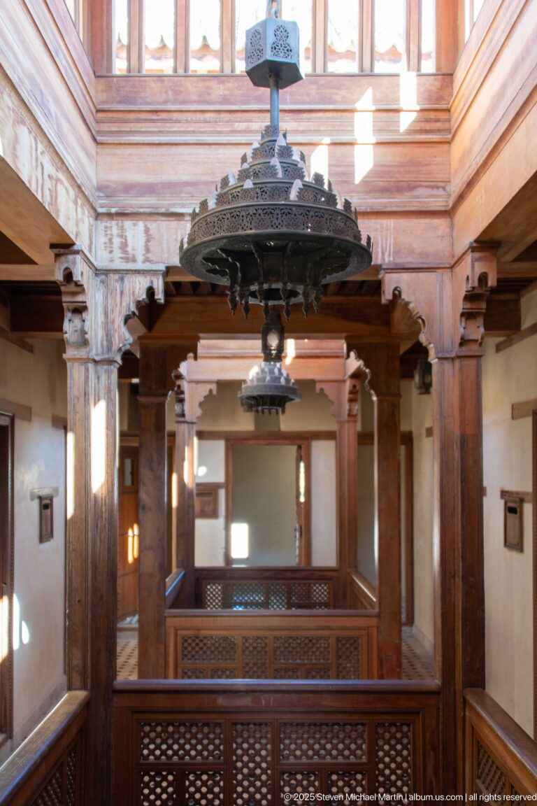 Large metal chandelier suspended over a cedar gallery with carved posts, lattice railings, and upper-level rooms.