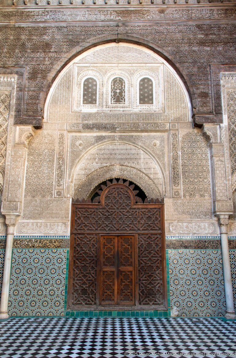 Carved wooden double door and screen beneath a monumental arch, surrounded by stucco carving and blue-green zellij.