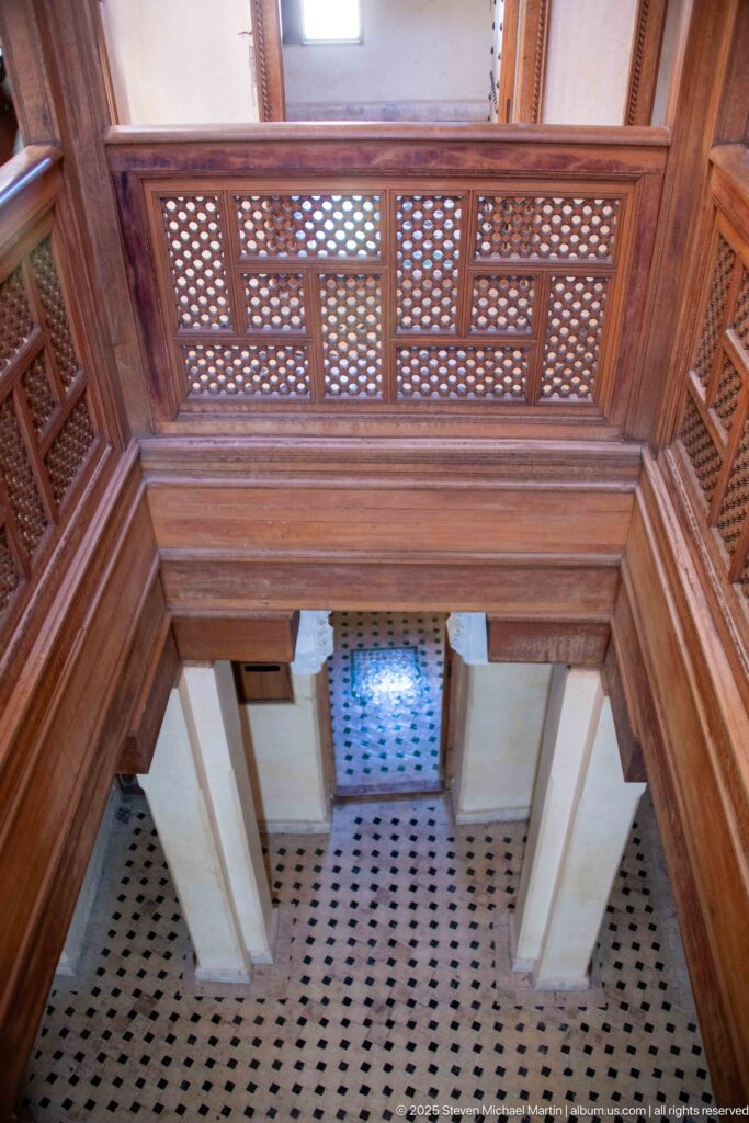 Upward diagonal view of cedar lattice panels, heavy beams, gallery rails, pale walls, and patterned floor.