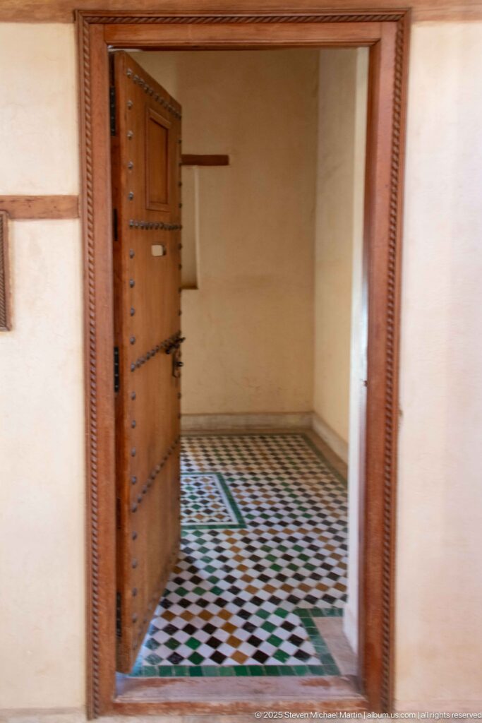 Open studded wooden door leading into a plain cream room with black, white, and green geometric floor tiles.