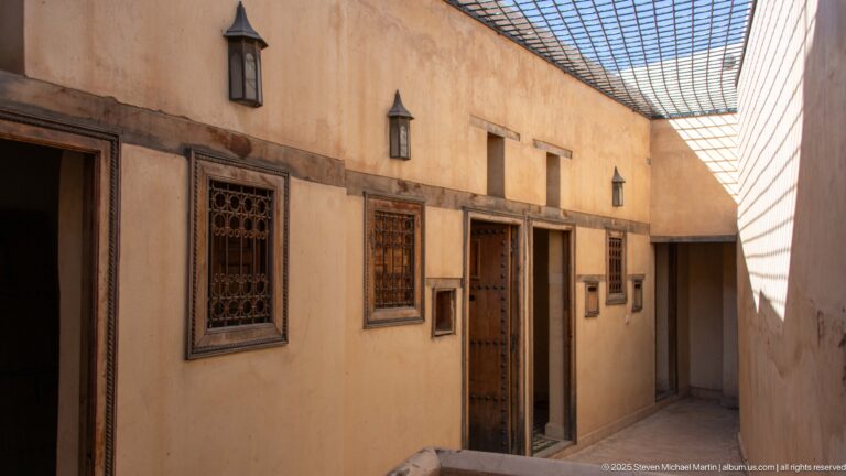 Narrow pale corridor with wooden doors, grilled windows, wall lanterns, and a mesh canopy casting striped shadows.