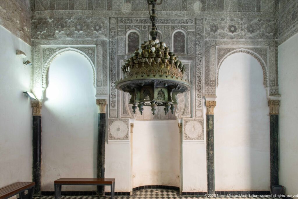 Prayer-hall interior with large hanging chandelier, carved stucco, tall arched recesses, marble columns, and central niche.