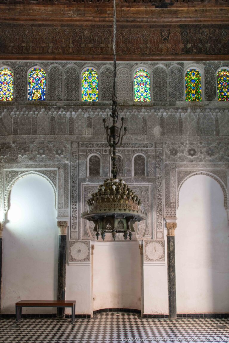 Attarine Madrasa prayer hall with glowing stained-glass windows above carved stucco, arched recesses, and a hanging chandelier.