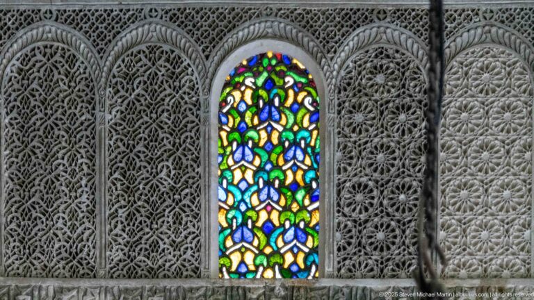 Narrow multicolored stained-glass window surrounded by dense carved stucco with geometric and vegetal patterns.