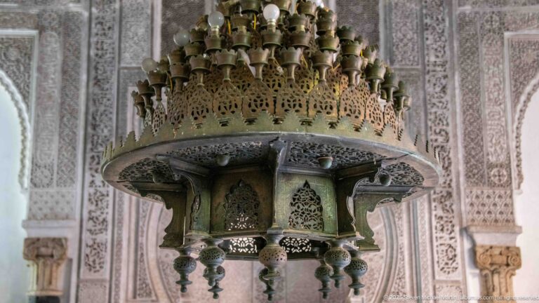 Close view of a large pierced metal chandelier with multiple lamp fittings suspended before carved stucco walls.