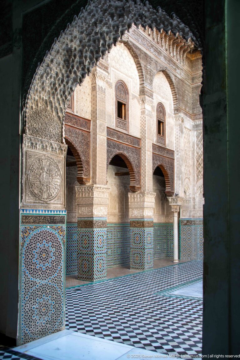 Sunlit Attarine Madrasa courtyard arcade seen through a shadowed arch, with upper windows, zellij, and black-and-white floor tiles.