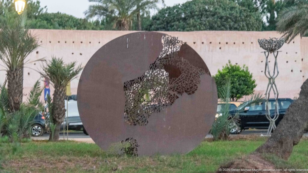 Rust-toned circular metal sculpture with a pierced openwork figure standing among palms and grass in Marrakech.