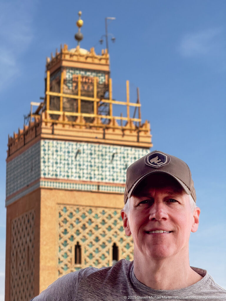 Smiling man wearing a baseball cap posed in front of the upper section of the Koutoubia minaret with scaffolding.