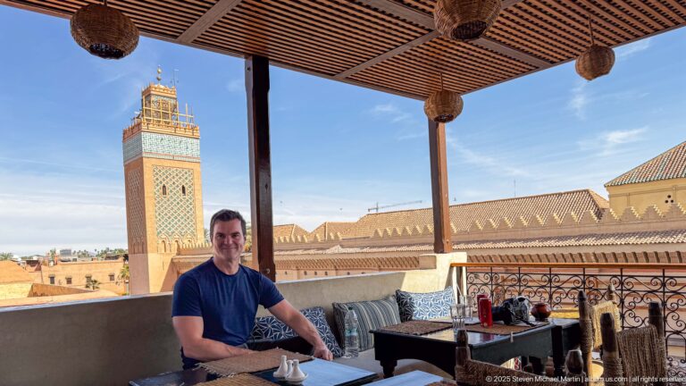 Man seated at a shaded rooftop terrace with cushions and table, overlooking the Koutoubia Mosque and minaret.