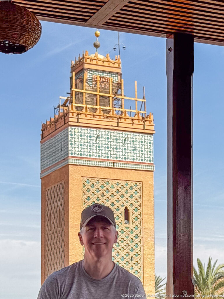 Smiling man in a cap beneath a rooftop canopy with the decorated Koutoubia minaret and scaffolding directly behind.