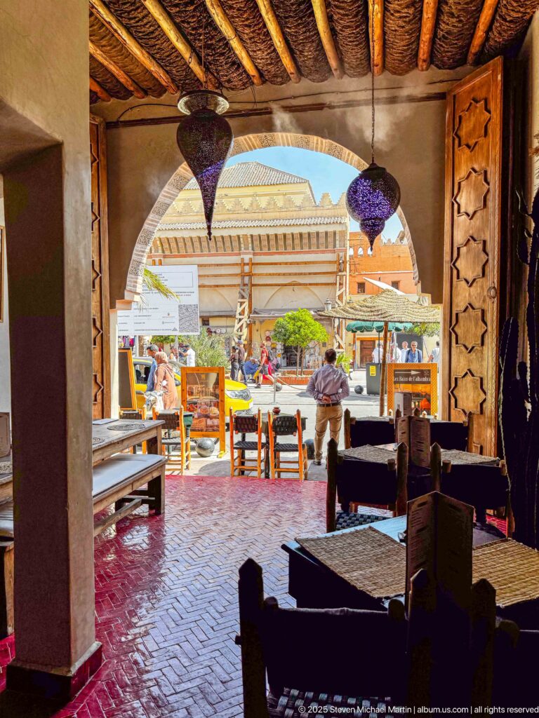 Shaded café interior with dark tables, hanging lamps, herringbone floor, and a large arch opening onto a bright Marrakech street.