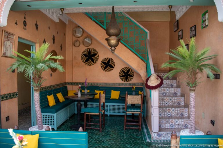 Turquoise Moroccan café interior with built-in seating, tiled stairs, potted palms, yellow cushions, and a large hanging lantern.