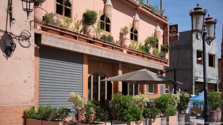 Pink multi-story restaurant or café frontage with geometric balcony panels, potted plants, umbrella, and street-level seating.