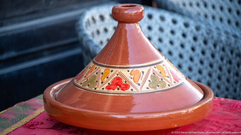 Glazed terracotta tagine with conical lid and painted red, green, yellow, and black decoration on a red tablecloth.