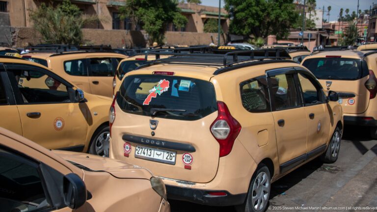 Several beige Marrakech petit taxis parked tightly together, showing roof racks, municipal emblems, and license plates.