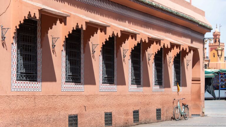 Pink façade with tall grilled windows, blue-and-white geometric tile borders, carved timber eaves, and a bicycle below.