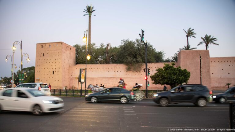 Pink-ochre fortified wall and square tower beside busy evening traffic on a Marrakech boulevard.