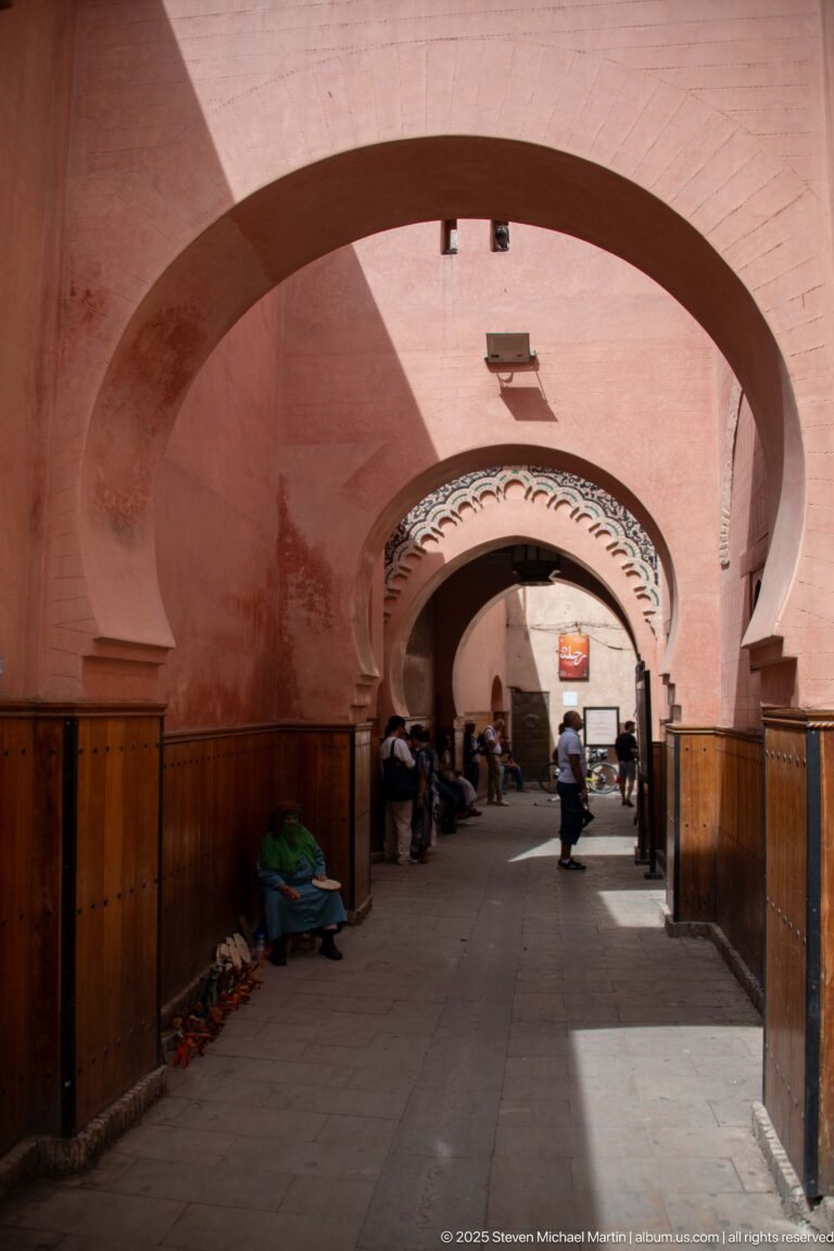 Long shaded Marrakech passage with repeating broad arches, pink plaster, wooden lower wall panels, vendors, and pedestrians.