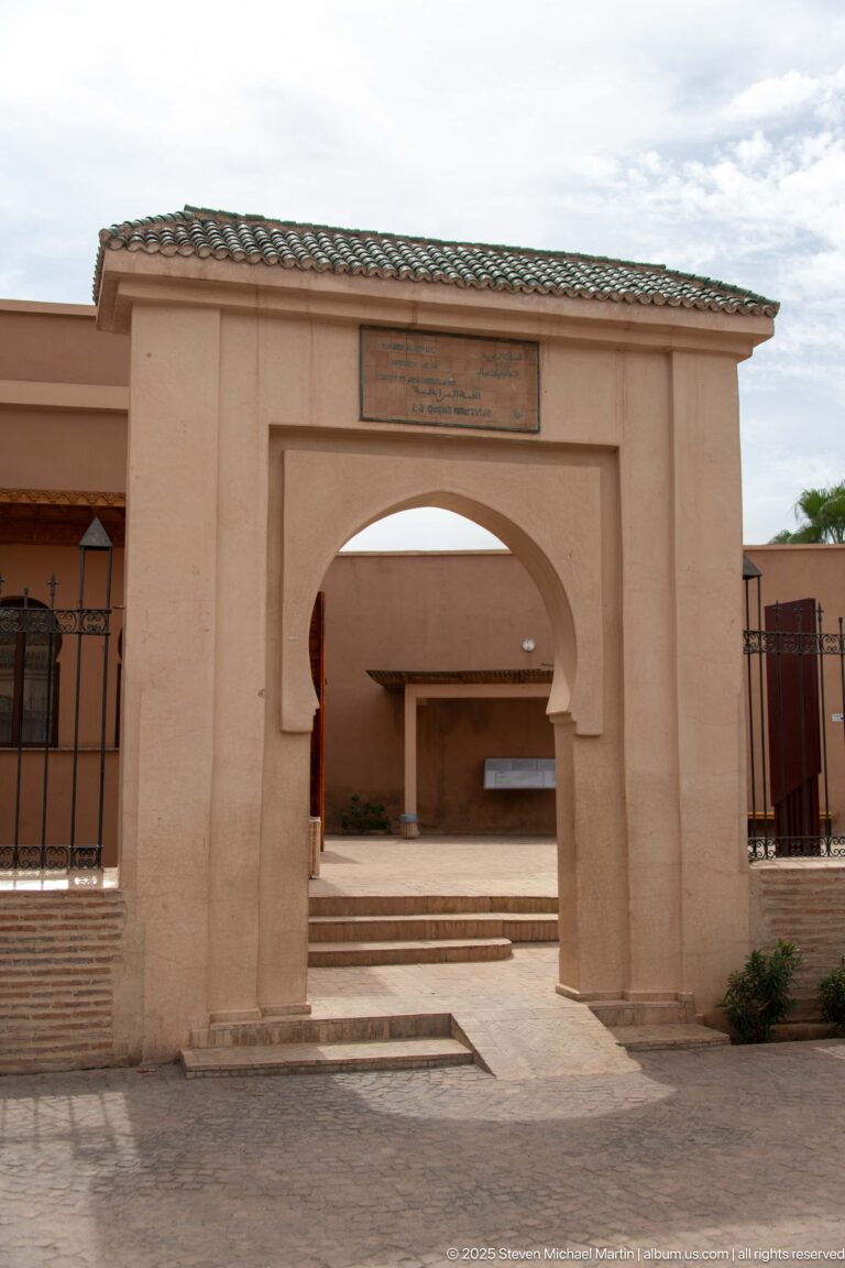 Pale entrance gateway with horseshoe arch and green-tiled roof leading into the Almoravid Qubba archaeological site.