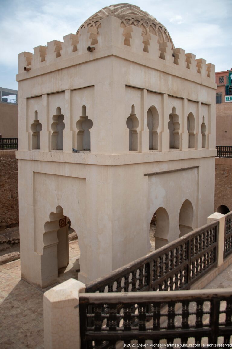 Oblique view of the Almoravid Qubba with repeated lobed openings, decorated dome, crenellations, and a descending balustraded stair.