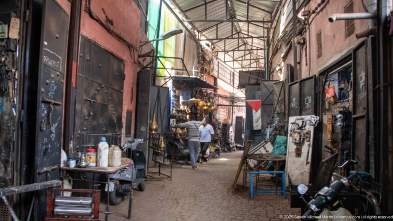 Covered Marrakech souk lane with metal roof, workshop doors, carts, stools, hanging goods, and pedestrians.