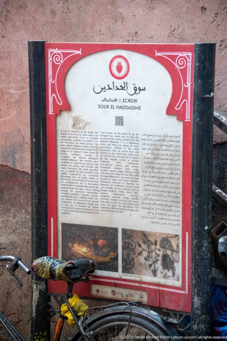 Red-bordered multilingual interpretive panel for Souk el Haddadine with Arabic, French, English text, historic images, and a bicycle below.