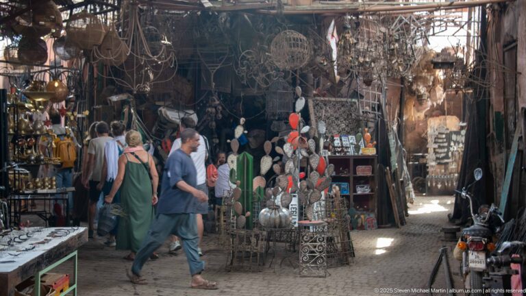 Pedestrians walking through Souk Haddadine among hanging metal lamps, mirrors, trays, wirework, tools, and workshop displays.