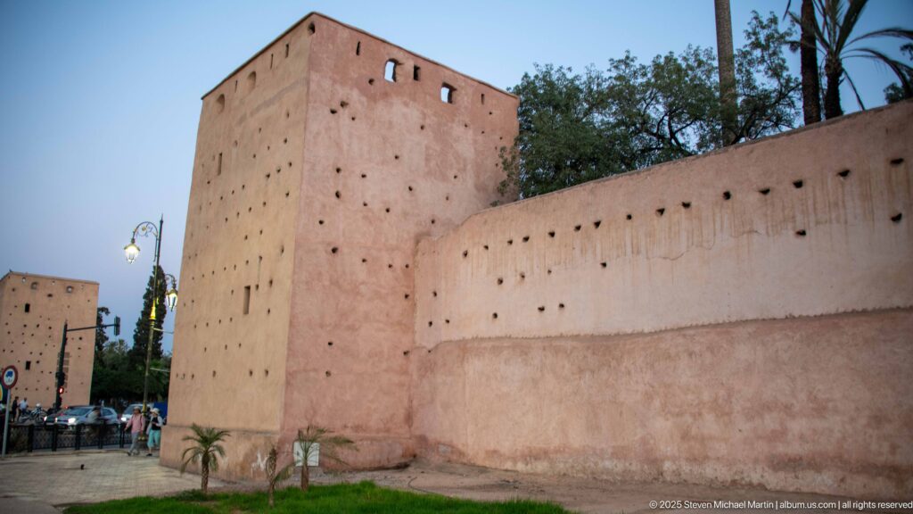 Square pink-ochre defensive tower joined to a long Marrakech city wall, with trees and palms nearby.