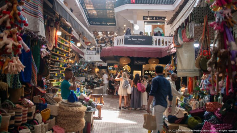 Busy covered Marrakech market with shoppers, white balconies, textiles, baskets, ceramics, and colorful souvenir displays.