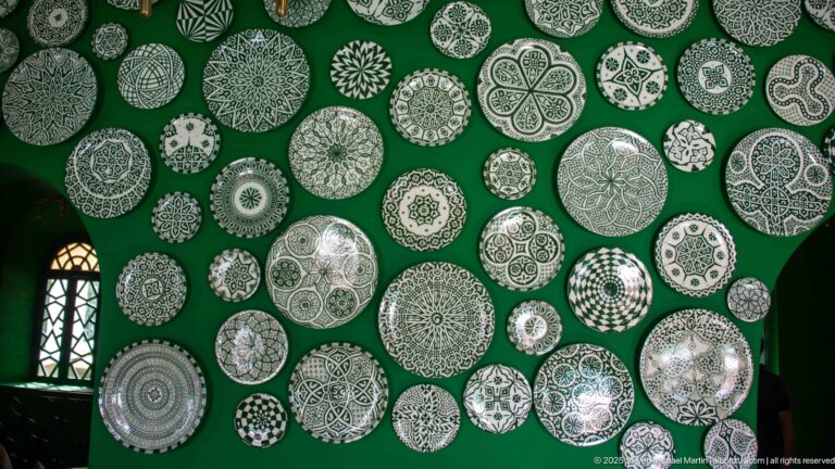 Dozens of patterned white ceramic plates arranged across a deep green wall in varied circular geometric and vegetal designs.